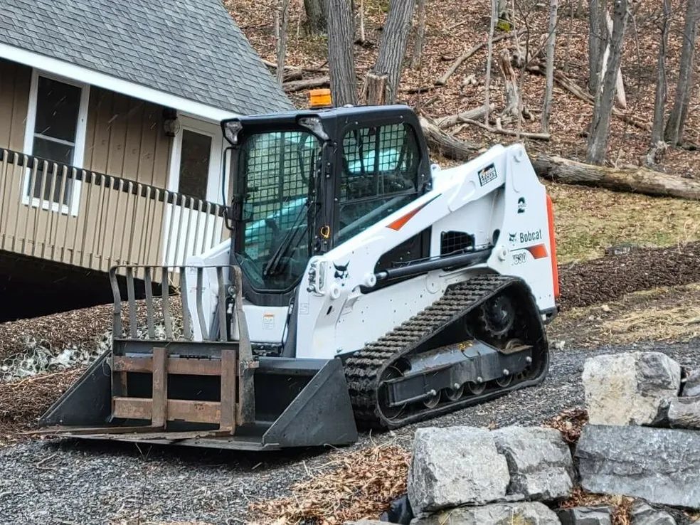 Bobcat compact track loader working beside a stacked-stone wall on a Rochester NY landscaping job